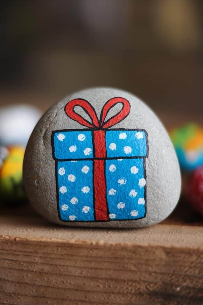A smooth gray rock with a hand-painted blue gift box design, featuring white polka dots and a red ribbon, rests on a wooden surface. Other colorful, blurred rocks are in the background.