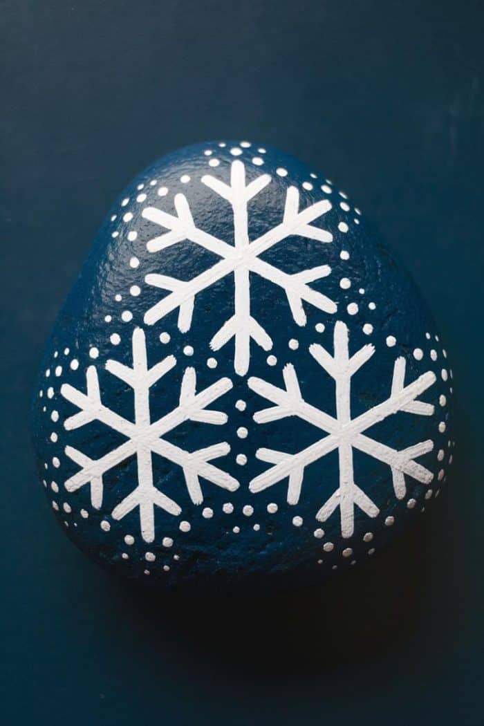 A smooth, dark blue stone is painted with three white snowflakes and dotted patterns. The background is a matching dark blue surface.