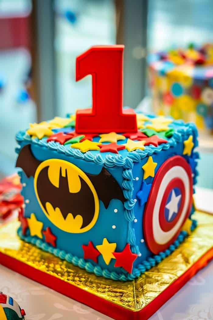 Colorful superhero-themed birthday cake in the shape of a cube, featuring a large red number "1" on top. The sides are decorated with Batman and Captain America logos, along with stars and swirls in a vibrant blue icing.