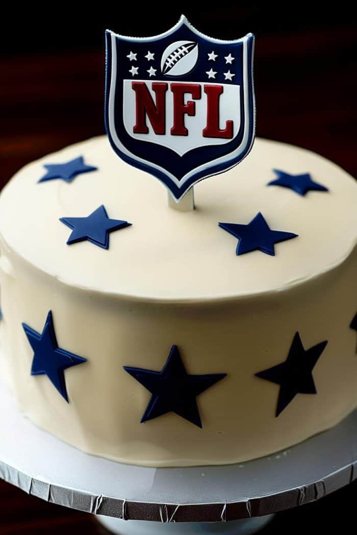 A round white cake decorated with dark blue stars. The cake is topped with a decoration featuring the NFL logo, which includes a football and shield design. The cake sits on a silver cake stand.