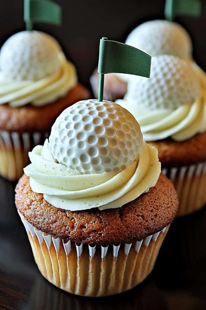 Cupcakes topped with white chocolate golf balls and small green flags. The cupcakes have swirls of vanilla frosting and are set against a dark background.