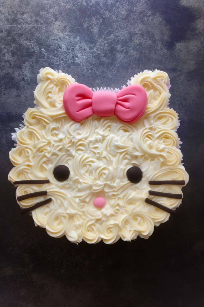 A pull-apart cake with cupcakes arranged to resemble Hello Kitty, with swirled white frosting for fur, chocolate pieces for whiskers and eyes, a pink candy nose, and a pink bow on top. The cake is set against a dark background.