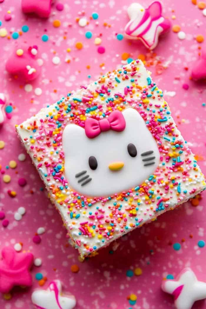A colorful square dessert with pink, yellow, and white sprinkles, featuring a Hello Kitty face with a pink bow. The dessert is set on a pink polka-dotted surface with star-shaped candies around it.