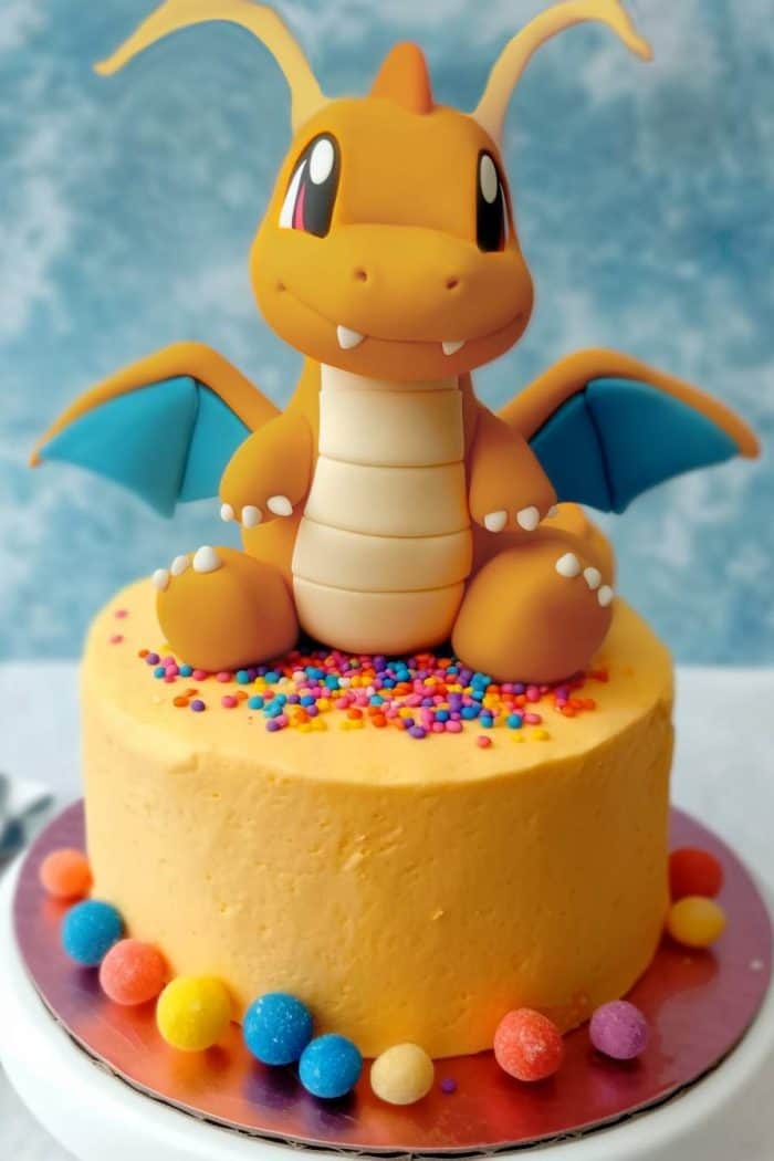 A round cake featuring a fondant figure of an orange, winged dragon with blue wings sitting on top. The cake is decorated with colorful sprinkles and surrounded by multicolored round candies on the base.
