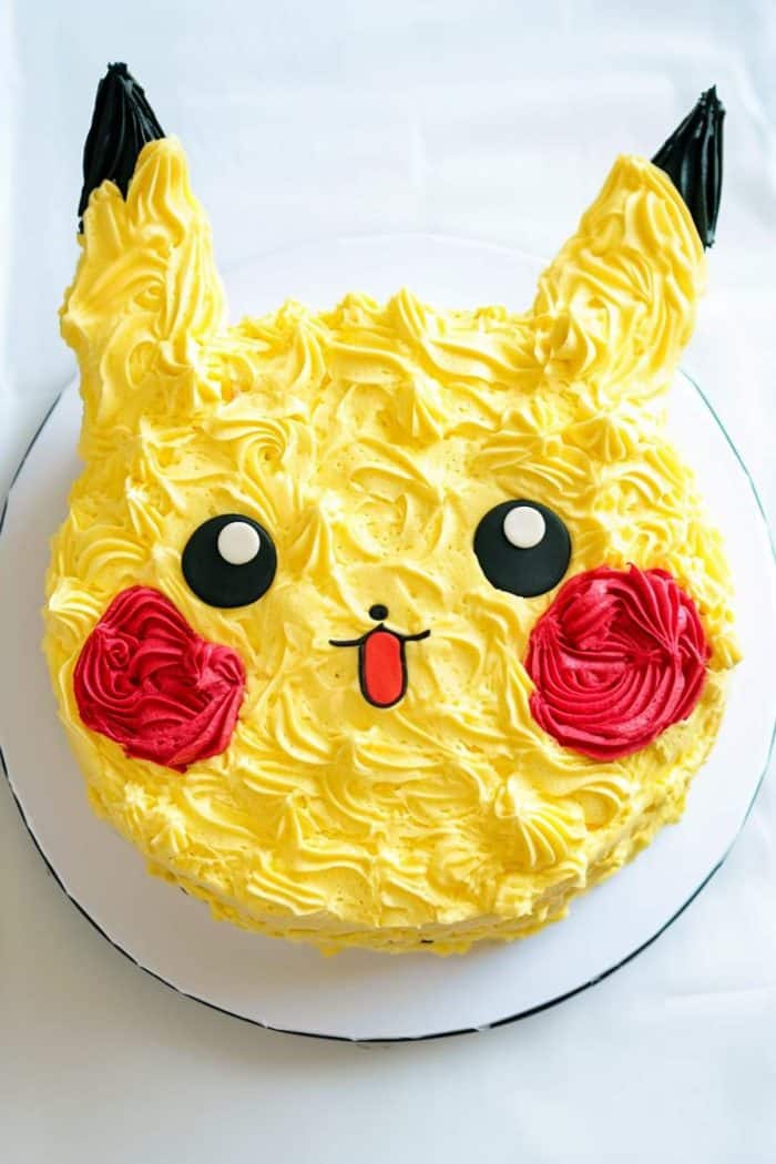 A cake decorated to resemble a cartoon character with yellow icing, large black eyes, red cheeks, and pointed ears. The mouth is open with a tiny red tongue. The cake is circular and placed on a white plate.