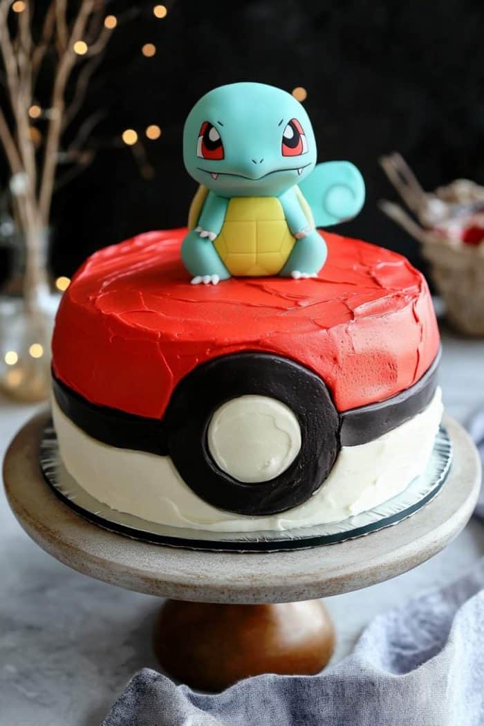 A Poké Ball-themed cake with smooth red, white, and black icing, topped with a fondant character resembling Squirtle, a blue turtle-like creature. The cake is displayed on a wooden cake stand against a blurred background.