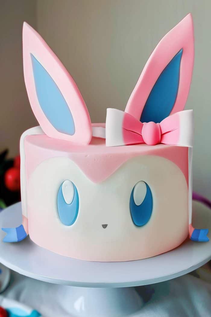 A round cake designed to resemble a pastel-colored animated character with large blue eyes, pink and blue ears, and a pink bow on top. The cake is displayed on a white stand.