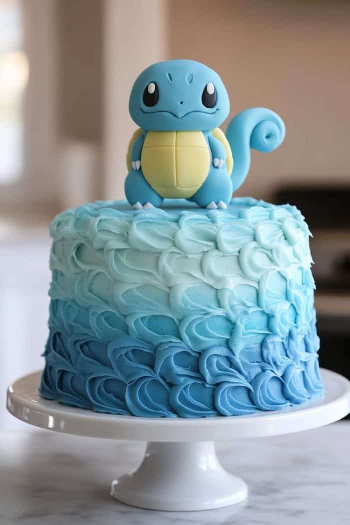 A cake with blue ombre frosting topped with a fondant figure of a cartoon turtle character, resembling a Pokémon, placed on a white cake stand.