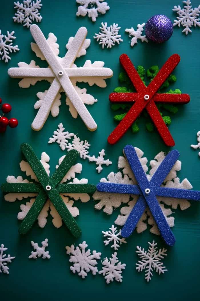 Crafted popsicle stick snowflakes in white, red, green, and blue on a teal surface, surrounded by small decorative snowflakes and baubles.