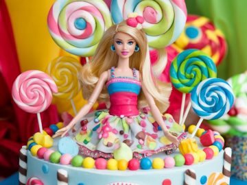 A colorful cake featuring a Barbie doll in the center, surrounded by large swirled lollipops and candies. The cake is decorated with bright colors, including pink, green, and blue, and has the word "Barbie" written in pink icing on the front.