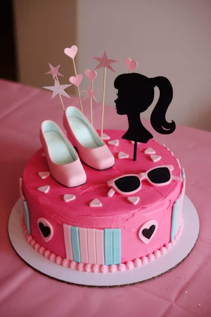 A pink cake adorned with heart decorations, a Barbie silhouette, pink shoes, and sunglasses. The cake is topped with star and heart-shaped toppers, set against a pink tablecloth.