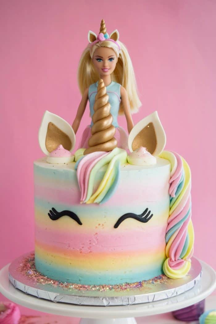 A colorful unicorn-themed cake topped with a doll. The cake features pastel rainbow frosting, unicorn ears, a golden horn, and a braid-like mane. The Barbie doll wears a unicorn headband and stands in the center. The background is pink.