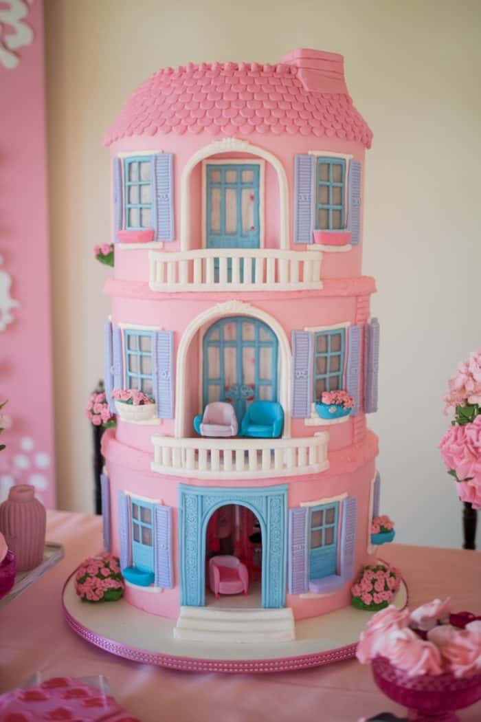 A multi-tiered Barbie cake designed to resemble the Barbie Dreamhouse with blue windows, shutters, and doors. Each tier has balconies decorated with pink flowers. The cake is detailed with textures mimicking roof shingles and architectural features.