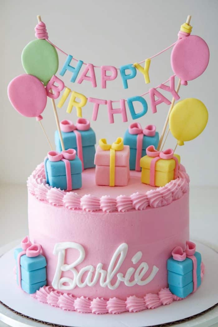 A pink birthday cake decorated with small colorful gift boxes and balloons on top. The cake has "Barbie" written in white icing and features a "Happy Birthday" banner made of letters strung across the top.