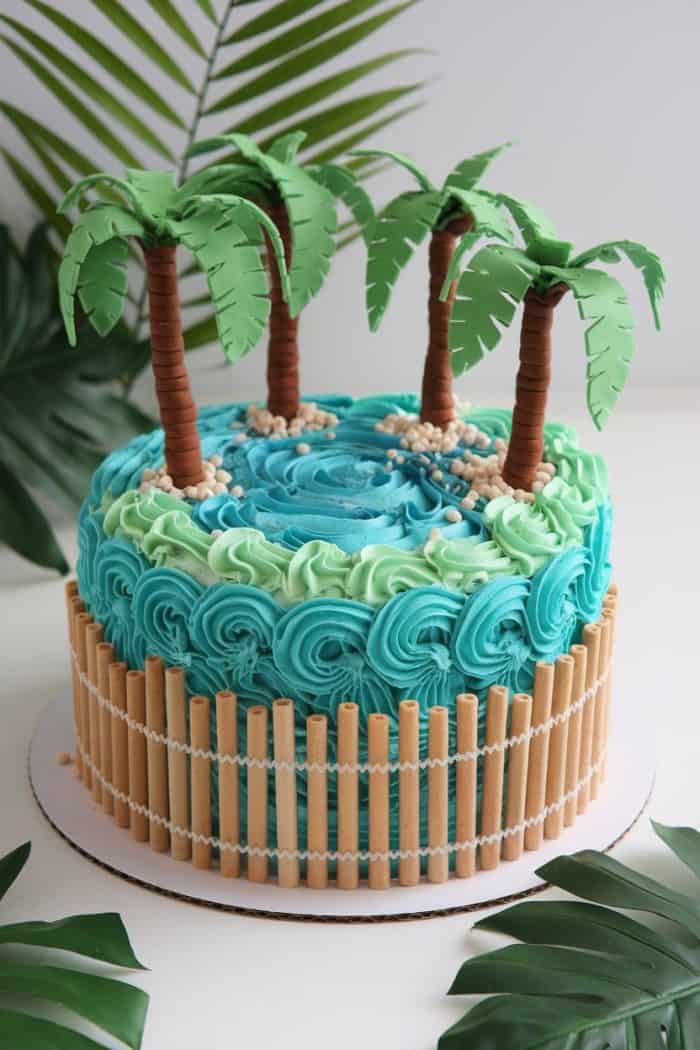 A cake decorated with blue and green frosting to resemble water and waves. It features three palm trees on top, with a border of wafer sticks around the sides. Palm leaves are placed in the background.