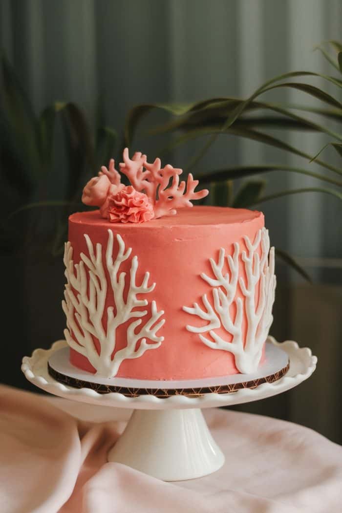 A pink cake adorned with white coral and pink flower decorations sits on a white, scalloped cake stand. Greenery in the background adds a natural touch to the elegant presentation.