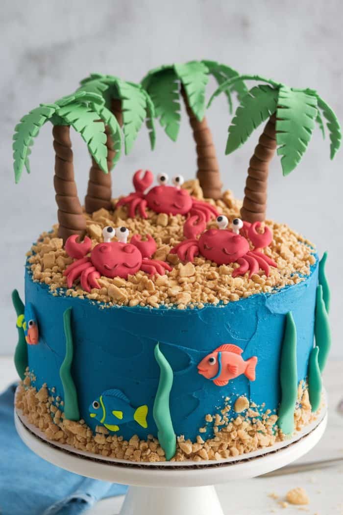 A beach-themed cake with blue icing, topped with three fondant crabs and palm trees. The sides feature fondant seaweed and fish, with crushed cookies resembling sand on top.