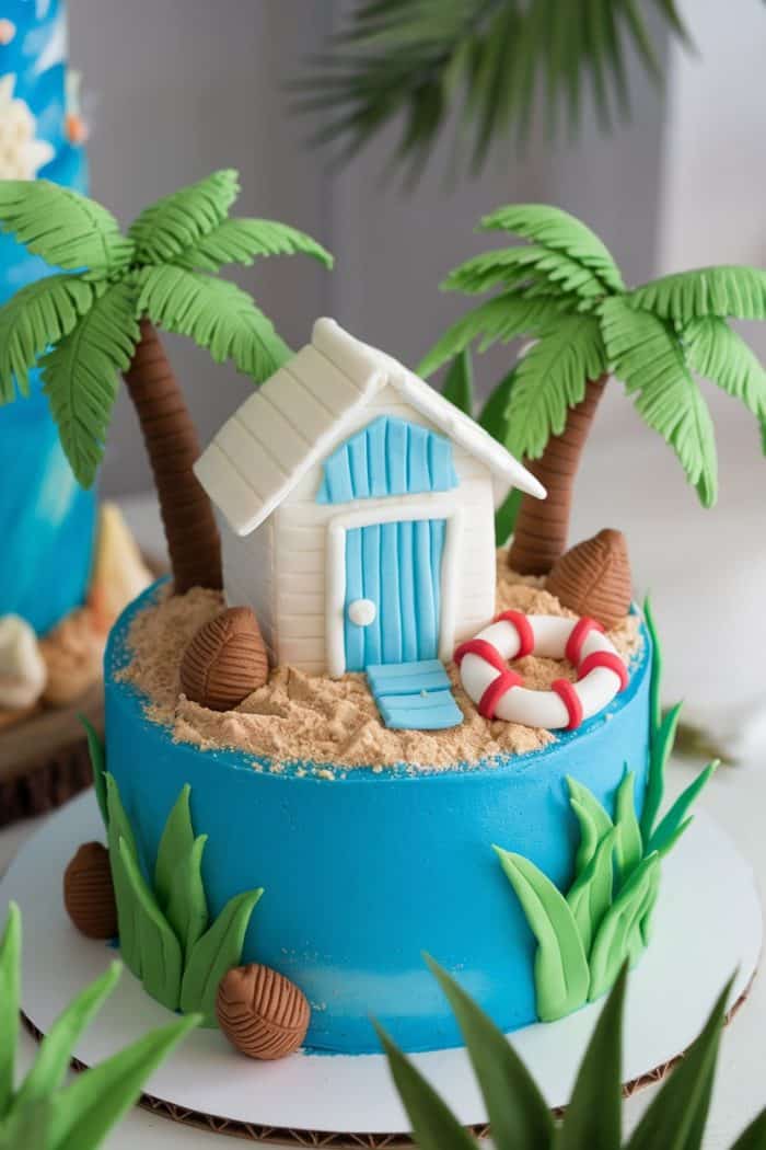 A beach-themed cake with a blue base, decorated with fondant palm trees, a striped beach hut, a lifebuoy, and scattered coconuts. The top resembles sandy terrain, adding to the coastal vibe.