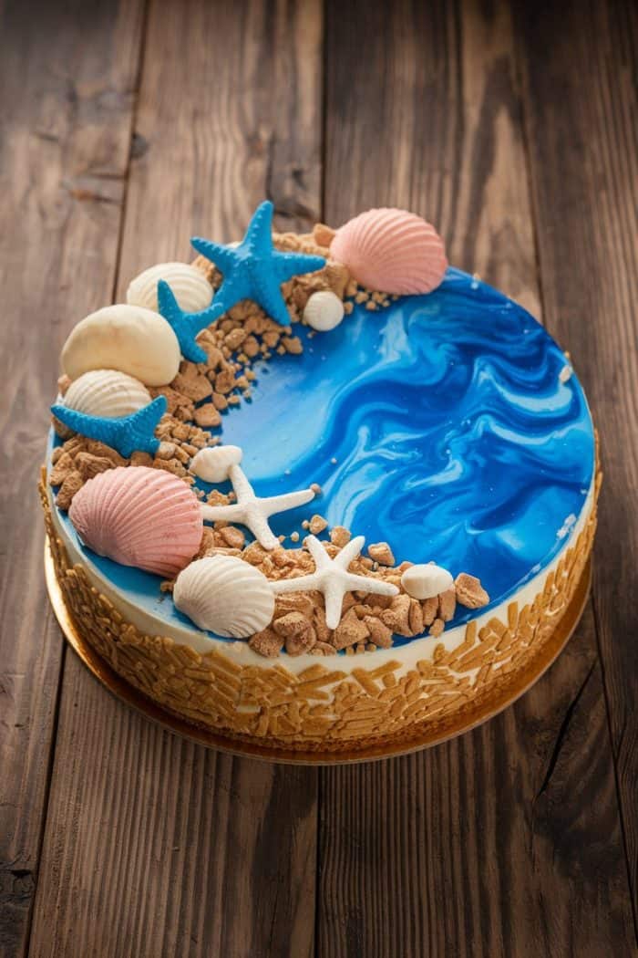 A beautifully decorated cake with a blue and white marbled fondant top resembling ocean waves. It features edible seashells, starfish, and crumbled cookie pieces, set on a wooden table.
