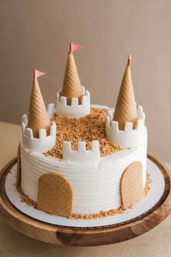 A castle-themed cake with white frosting and ice cream cones as towers, featuring small flags on top. Crushed cookies form a moat, and wafer panels resemble gates on the round cake. It's placed on a wooden stand.
