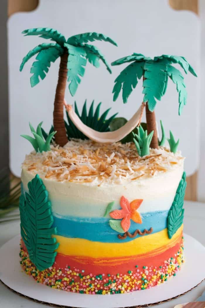A colorful tropical-themed cake with icing layers in blue, orange, and yellow. It features edible palm trees, a hammock, leaves, and a flower on top. The base is decorated with sprinkles.
