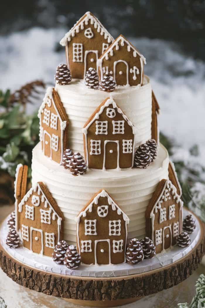 A two-tiered cake decorated with white icing and topped with small gingerbread houses and pine cones. The cake sits on a wooden base against a snowy background, creating a festive, wintry scene.