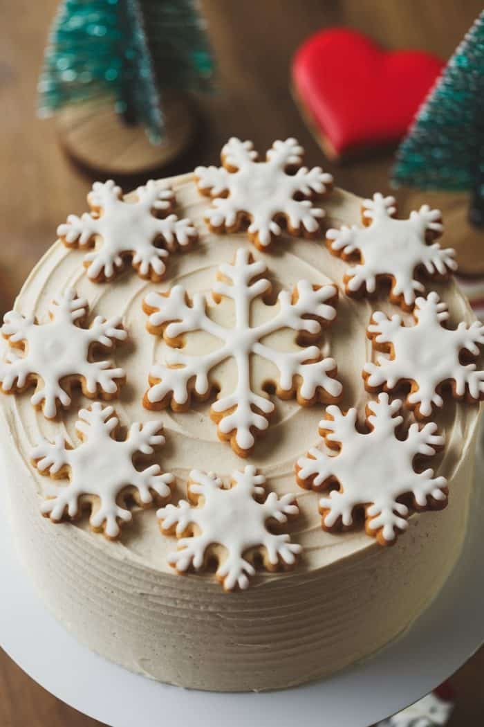 A frosted cake is decorated with white snowflake-shaped cookies. In the background, there are miniature Christmas trees and a red heart ornament on a wooden surface.