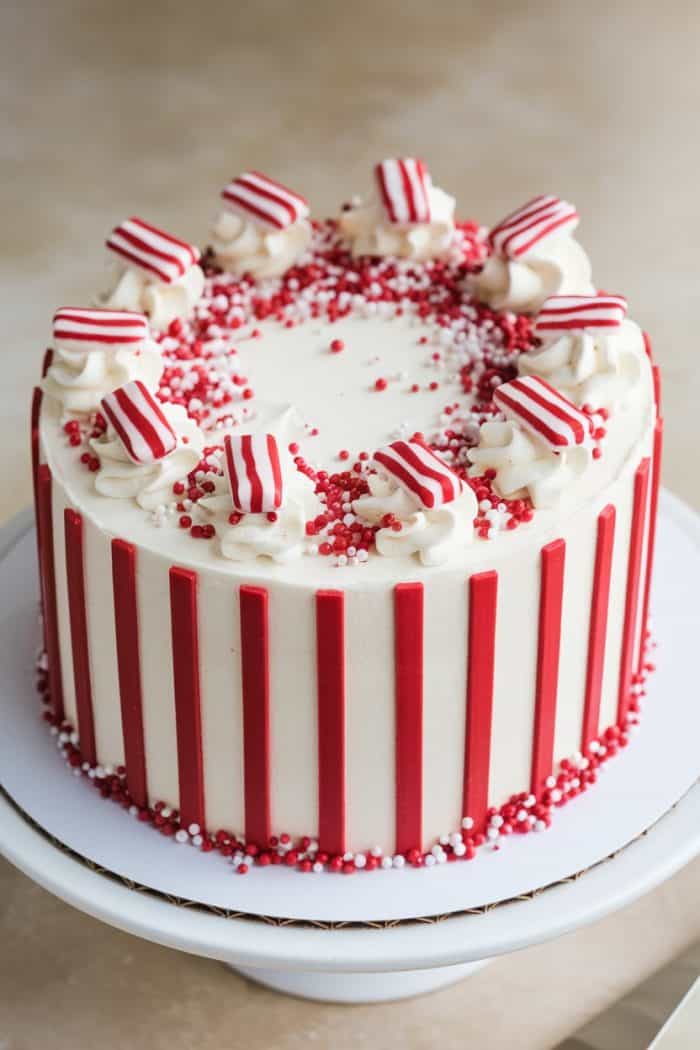 A round cake with red and white vertical stripes. The top is decorated with red and white sprinkles and striped red and white candy pieces. It sits on a white cake stand.