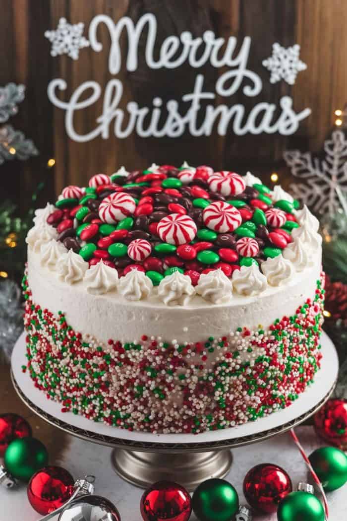 A festive Christmas cake adorned with red and green candies and swirls, topped with a Merry Christmas sign. The cake is surrounded by red and green ornaments and holiday decorations on a wooden background.