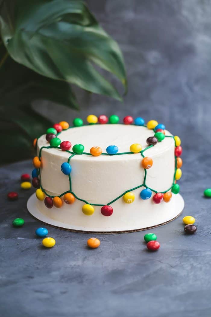 A white frosted cake is decorated with colorful round candies arranged to resemble a string of lights. Some candies are scattered at the base. A green leaf is partially visible in the background.