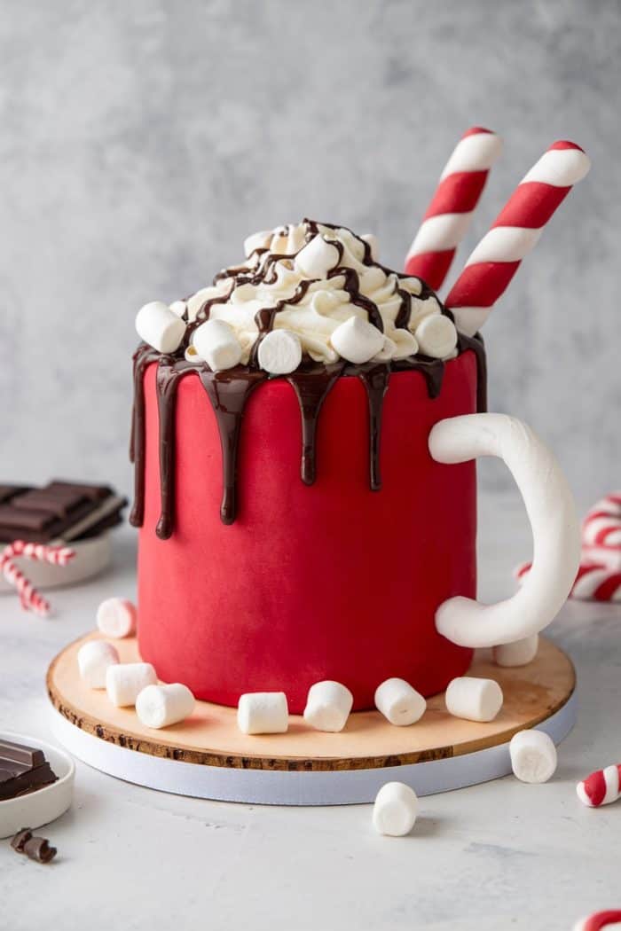 A cake shaped like a red mug, topped with whipped cream, chocolate drizzle, and mini marshmallows. Two red and white striped straws are inserted on top. The cake sits on a wooden base, surrounded by more marshmallows.