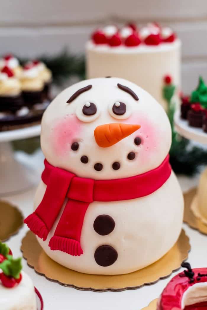 A festive cake shaped like a cute snowman, with a fondant carrot nose, pink cheeks, chocolate eyes, and red scarf and buttons. It is surrounded by other small cakes and holiday decorations.