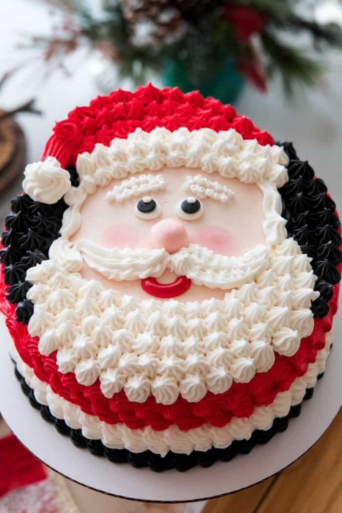 A festive cake decorated to look like Santa Claus, featuring a red hat, white beard, bushy eyebrows, and rosy cheeks. The cake is adorned with red, white, and black icing, resembling Santa's traditional outfit.