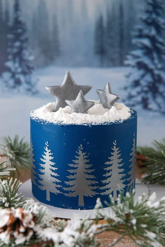 A blue cake decorated with white stencil pine trees, topped with silver star-shaped decorations and powdered sugar to resemble snow, is surrounded by snowy pine branches. The background features a frosty forest scene.
