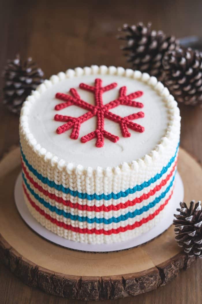 A decorated cake with white frosting and a red snowflake design on top. The sides feature a woven pattern in red, white, and blue. It is placed on a wooden slab and surrounded by pinecones.