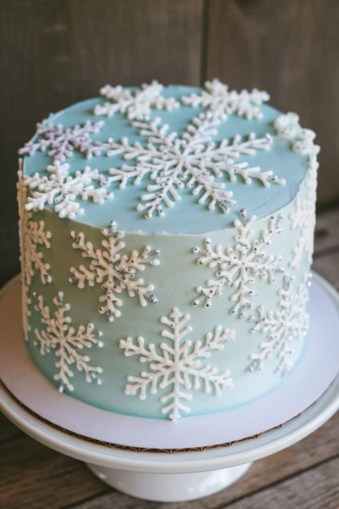 A round cake with light blue icing adorned with white snowflake decorations and small silver beads. It sits on a white cake stand, with a rustic wooden background.