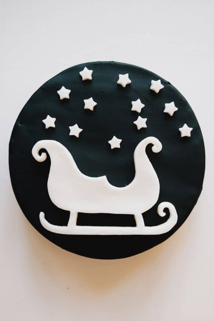 A round, black frosted cake with a white fondant silhouette of a sleigh and scattered white stars on top.