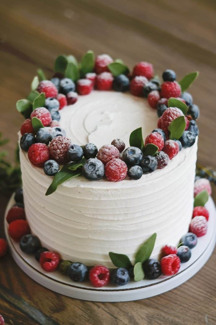 A frosted white cake garnished with a ring of fresh raspberries, blueberries, and green leaves on top. More berries and leaves decorate the base. The cake sits on a wooden table.