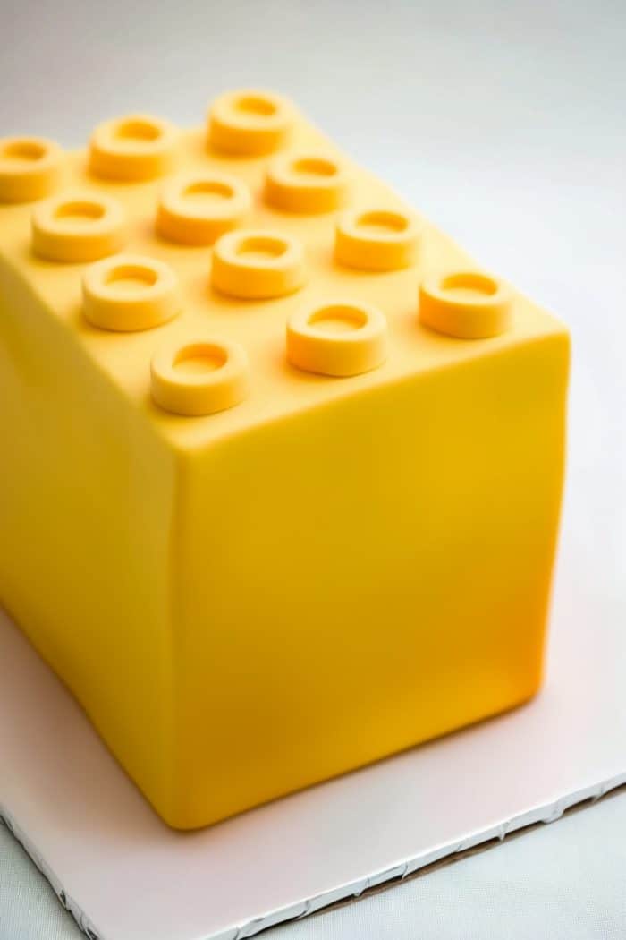 A rectangular cake designed to resemble a large yellow plastic building block with raised circular knobs on top, set on a white surface.