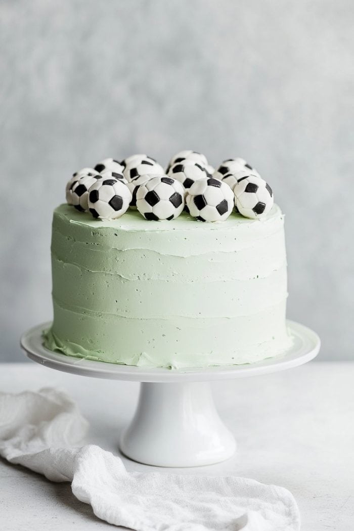 A light green frosted cake on a white stand, topped with small black and white soccer ball decorations, sits on a white surface with a soft gray background. A white cloth is draped nearby.
