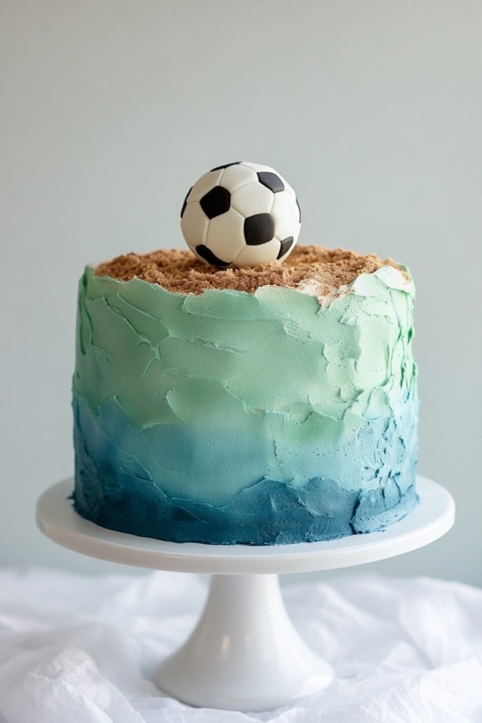 A round cake with blue and green icing resembling waves and a sandy top. A soccer ball decorates the top. The cake sits on a white cake stand against a light background.
