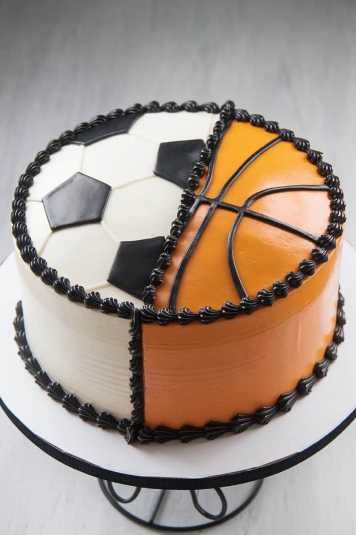 A round cake decorated to resemble a soccer ball on one half and a basketball on the other. The cake features black icing trim and is placed on a metal cake stand.
