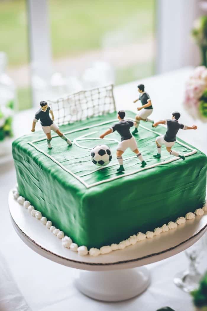 A square cake decorated as a soccer field, topped with green icing. Four soccer player figurines and a ball are positioned on top. The cake sits on a white stand with a window view in the background.