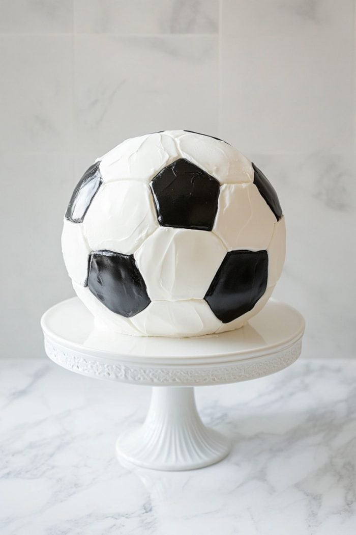 A cake designed to look like a classic black and white soccer ball is displayed on a white pedestal cake stand, set against a light marble background.