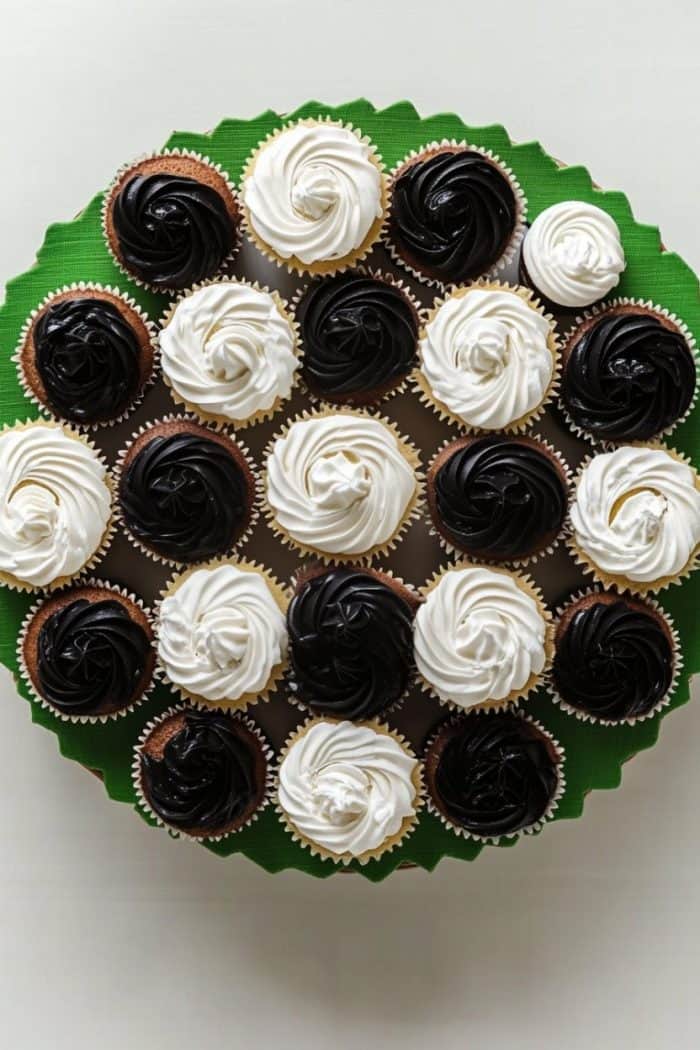 A circular arrangement of cupcakes with alternating black and white frosting on a green background. The cupcakes are neatly aligned, creating a visually appealing pattern.