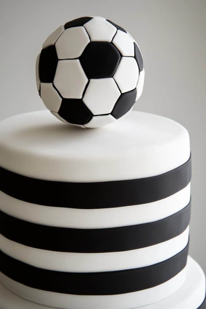 A two-tiered cake decorated with black and white fondant stripes. A realistic soccer ball made of fondant sits on top, complementing the cake's sporty theme. The background is a simple, light gray color.