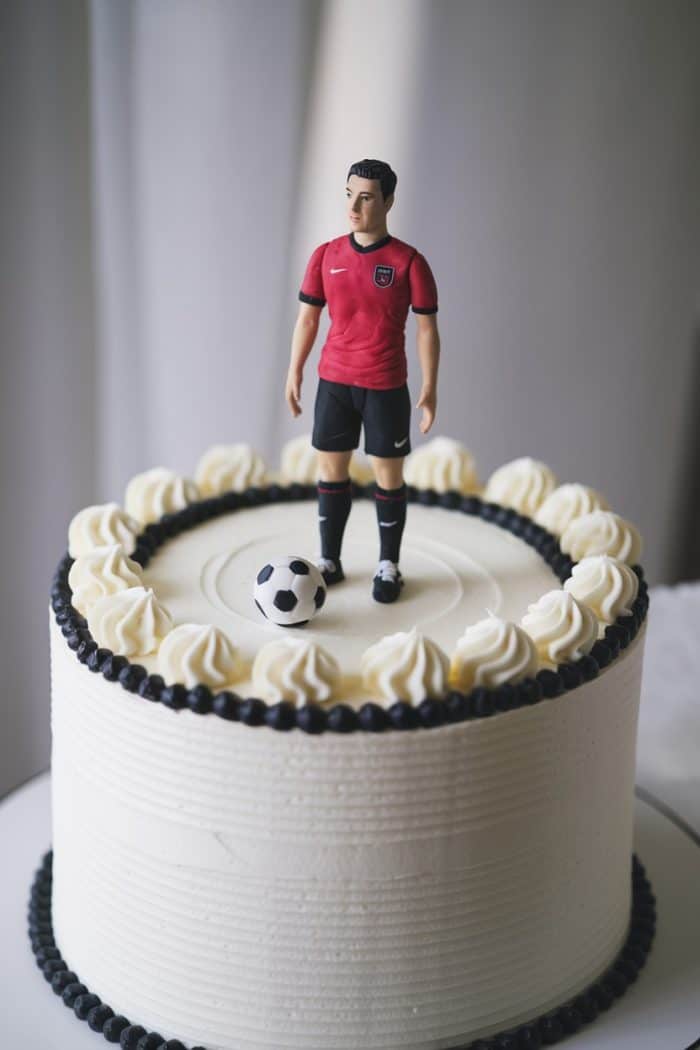 A round cake topped with a figurine of a soccer player in a red jersey and black shorts. The cake is decorated with white frosting and has a black and white pattern along the edge. A small soccer ball sits near the figurine's feet.