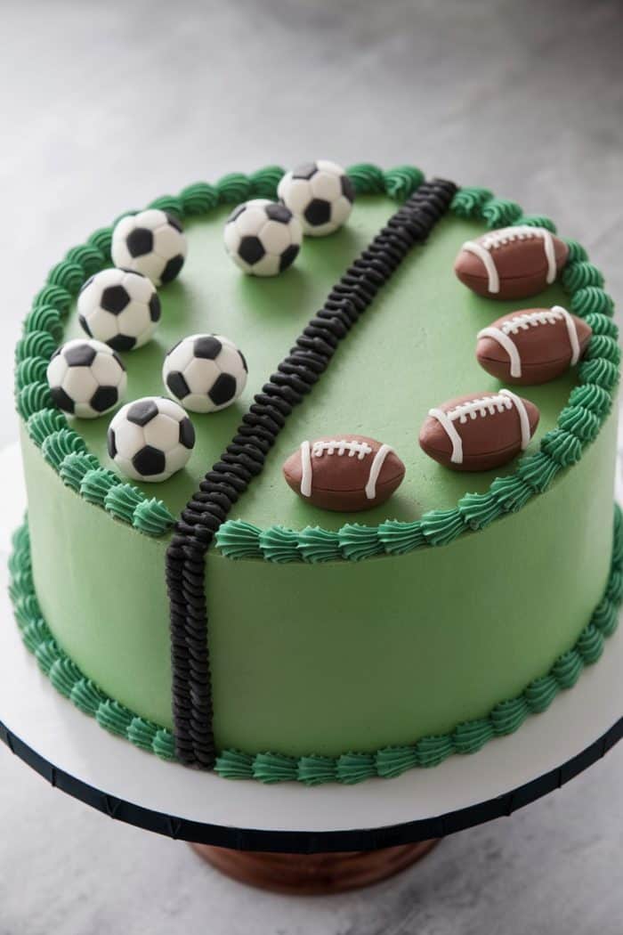 A round cake with green icing, decorated with small edible soccer balls on the left and footballs on the right. A black icing line divides the cake in half, surrounded by a green icing border at the base and top edge.