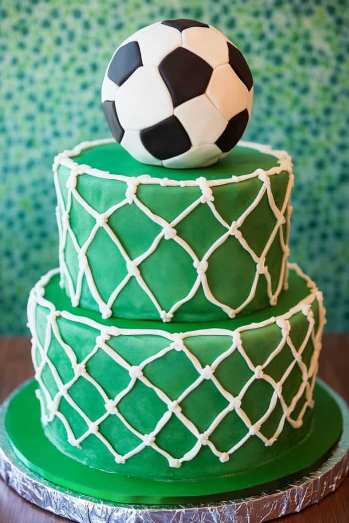 A two-tiered green cake decorated with white crisscross patterns resembling a soccer net. A soccer ball made of fondant is placed on top as a cake topper. The background features a textured green pattern.