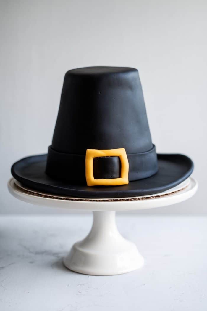 A cake designed to look like a black pilgrim hat with a yellow buckle sits on a white cake stand against a light gray background.